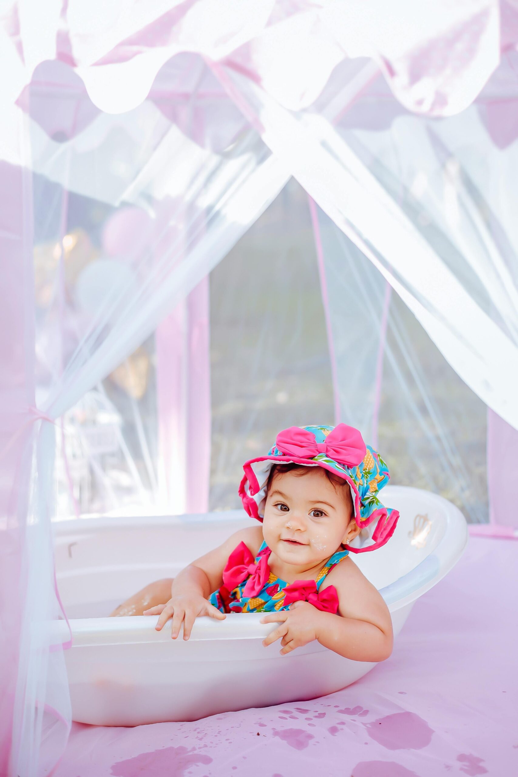 Cute baby wearing floral hat and swimsuit in pink tent plays in a bathtub, exuding pure joy.